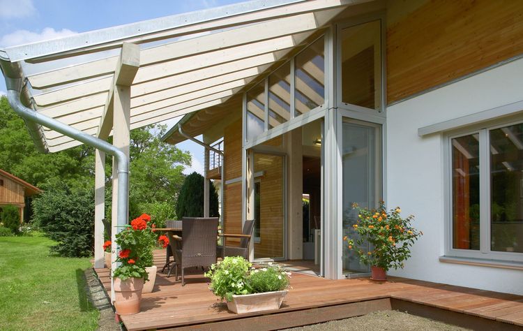 Die überdachte Terrasse des Holzhauses im Landhausstil mit geschickt eingesetzte Mischfassade aus mineralischem Außenputz und Lärchenholz und Schiebläden, die elektrisch verstellbar sind