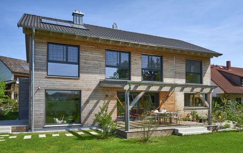 Die Terrasse des modernen Holzhauses im Landhausstil mit Holzfassade aus Lärchenholzbohlen und Doppelcarport