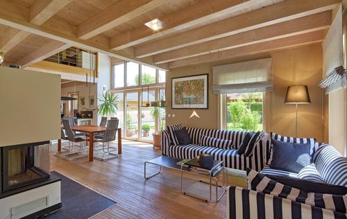 Das Wohnzimmer des Holzhauses im Landhausstil mit Lärchenfassade, großer Glasfront und moderner Technik mit Kachelofen und gestreifter Couch
