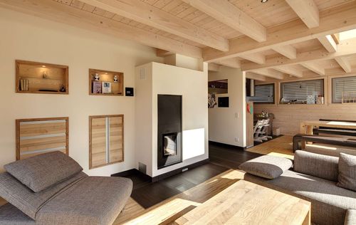 Das Wohnzimmer mit Kachelofen des Holzhauses im Landhausstil mit durchgängiger Holzfassade und großer Glasfront
